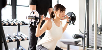 Asian woman doing barbell squats with the assistance of a trainer